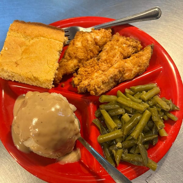 A red divided plate holds fried chicken tenders, cornbread, mashed potatoes with gravy, and green beans.