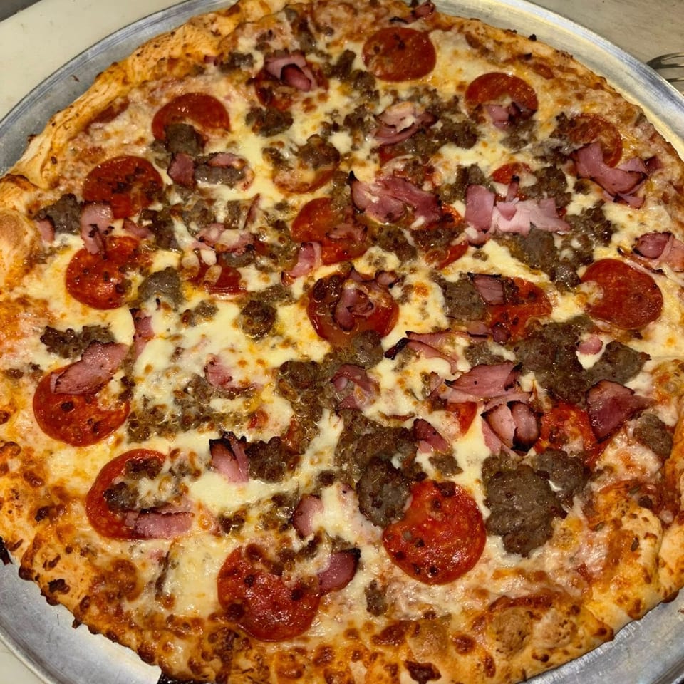 A large, round meat lover's pizza with pepperoni, sausage, and ham on a metal tray.