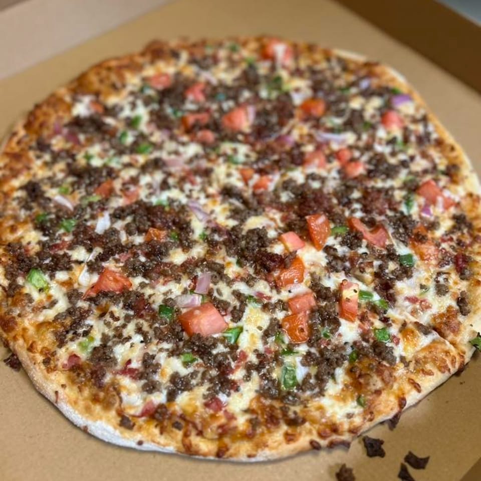 A whole pizza with ground beef, diced tomatoes, green peppers, and onions in a cardboard box.