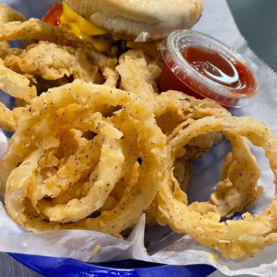 Crispy golden onion rings with black pepper, a burger, and a side of ketchup in a blue basket.