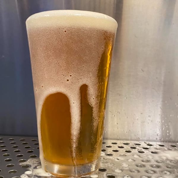 A pint glass of golden beer with a thick head of foam, sitting on a metal grate.