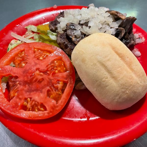 A deconstructed burger on a red plate with a tomato slice, bun, patty, onions, and mushrooms.