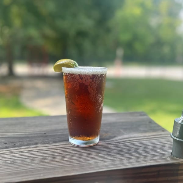 A cold, dark beverage with a lime wedge and frothy head sits on a wooden table outdoors.
