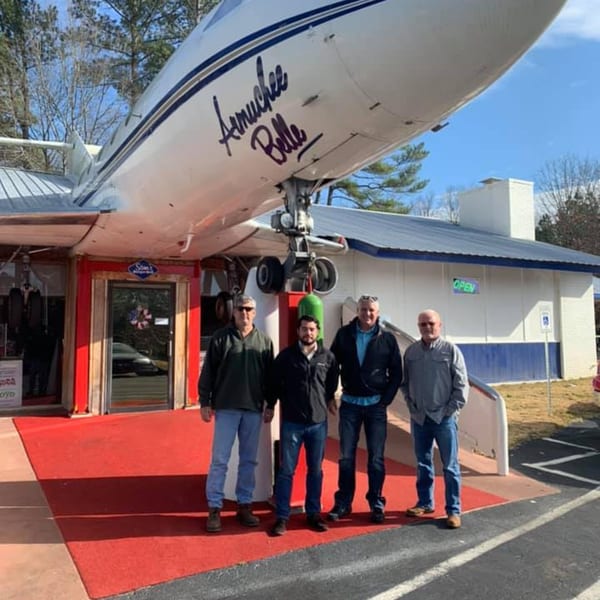 Four men stand on a red carpet in front of Sam's Burger Deli, housed inside a large airplane.